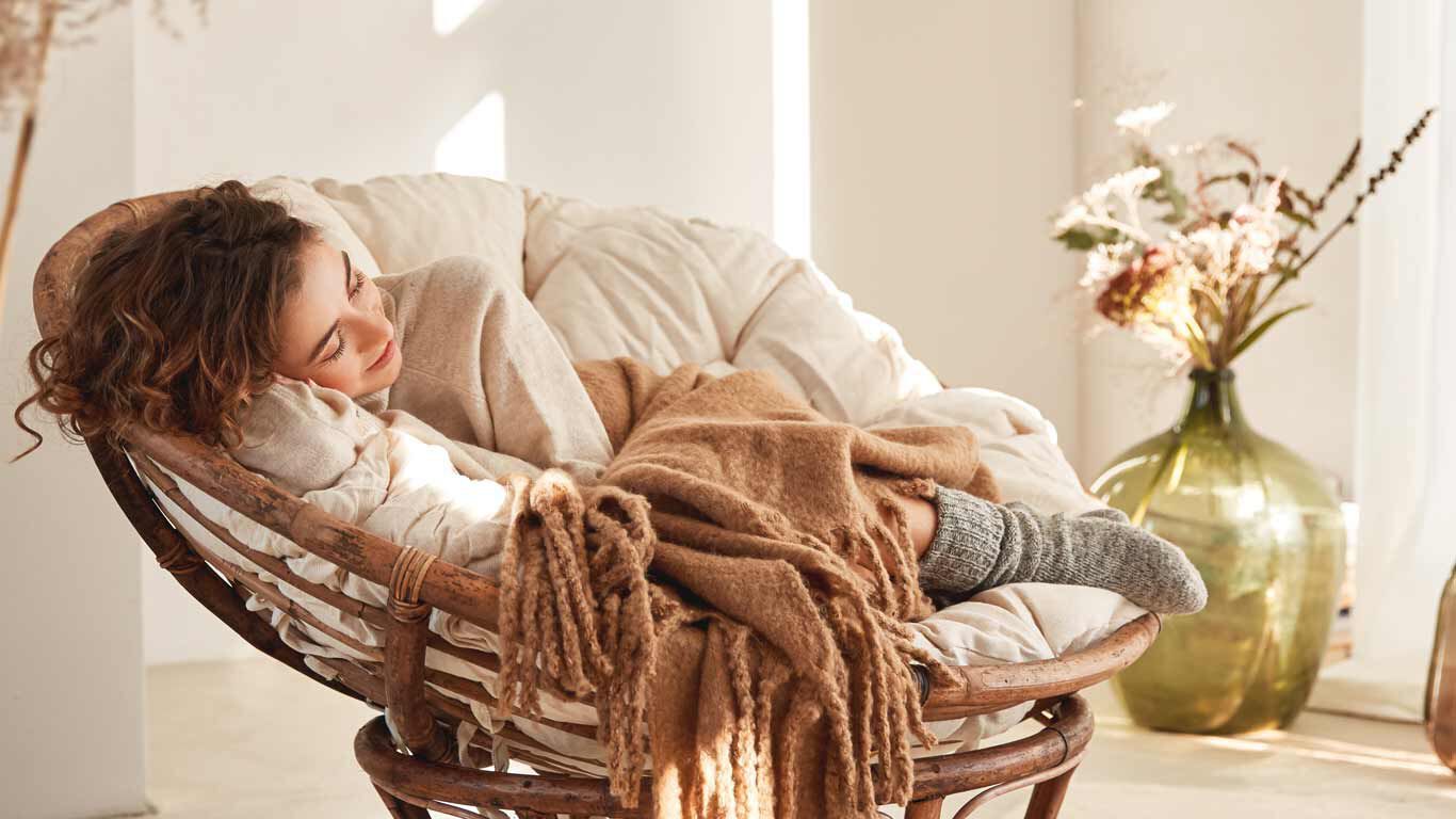 Een vrouw zit ontspannen in een ronde houten fauteuil met een deken over zich heen. 